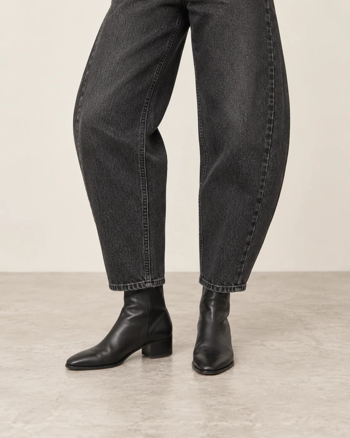 Washed-black barrel jeans taper around slim black ankle-boot shafts, covering the ankles while the hems hang freely outside the boots.