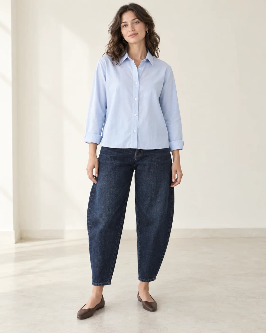A short pale-blue boxy shirt covers the waistband of dark-indigo barrel jeans, with the curved legs and chocolate almond-toe flats visible.